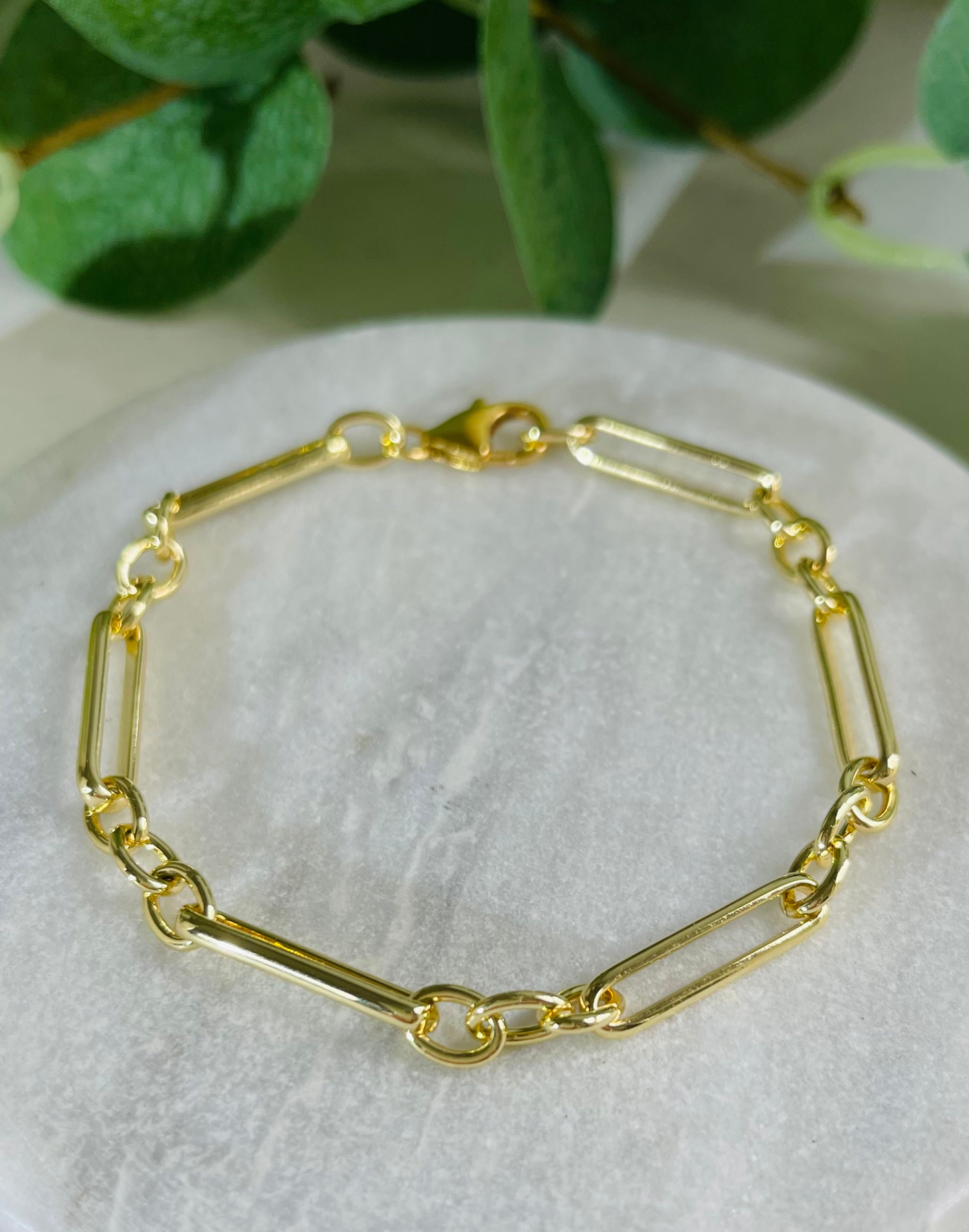Gold bracelet on a white surface with green leaves in the background
