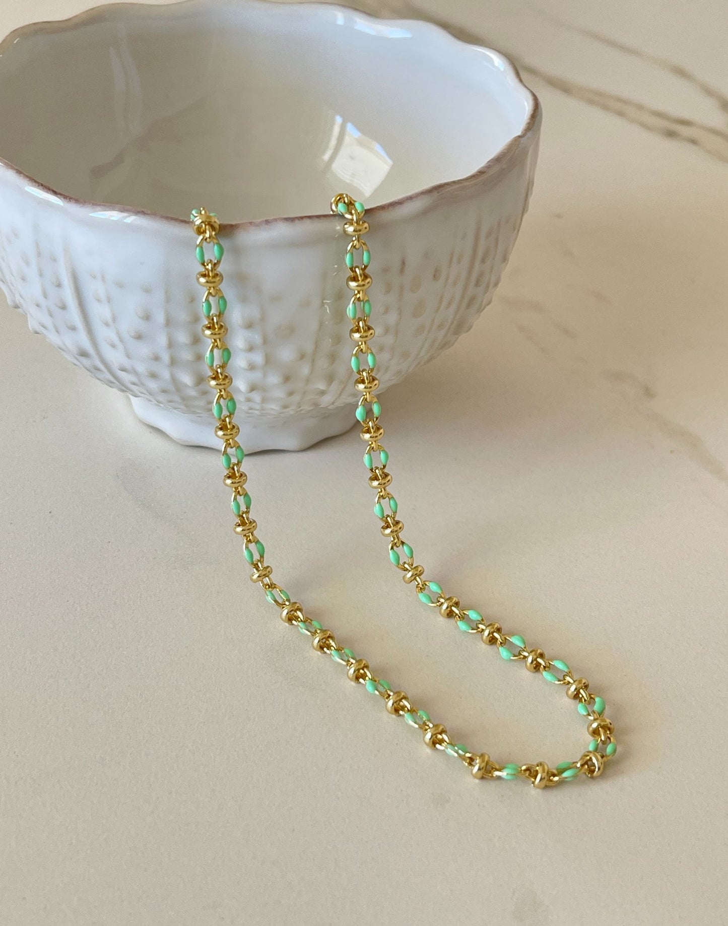 Gold and green enamel necklace on a light surface with a white textured bowl in the background