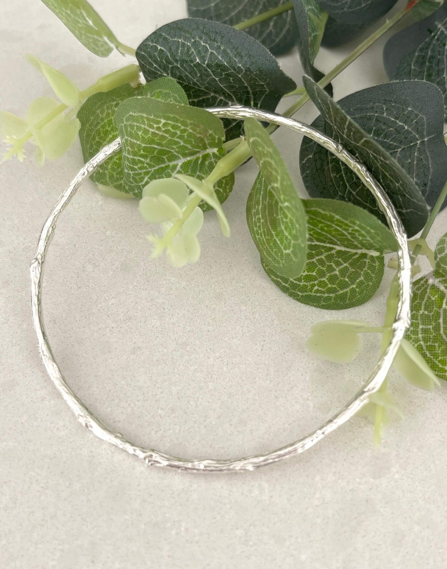 Twig silver bangle on a light grey background with green leaves
