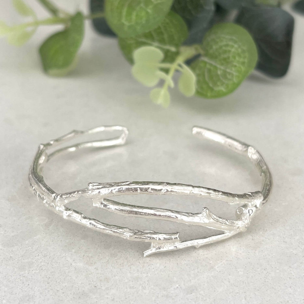 Silver Twig Cuff bracelet on a white 
background with green leaves in the corner