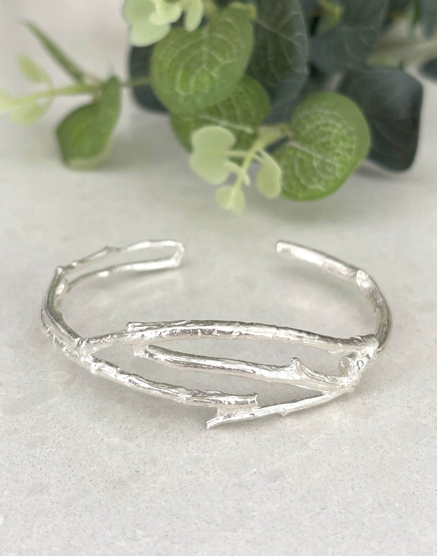 Silver Twig Cuff bracelet on a white 
background with green leaves in the corner